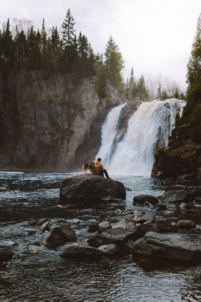 白天，一名身穿棕色外套的妇女坐在瀑布附近的岩石上